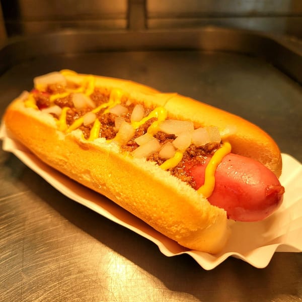 A US Coney and Cone hot dog with chili, mustard, and diced onions in a paper tray.