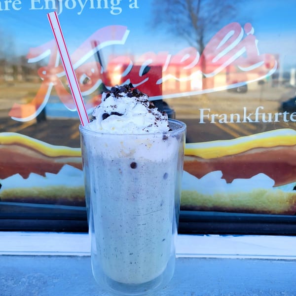 Oreo milkshake with whipped cream and a red-striped straw, in front of a window with the US Coney and Cone logo.