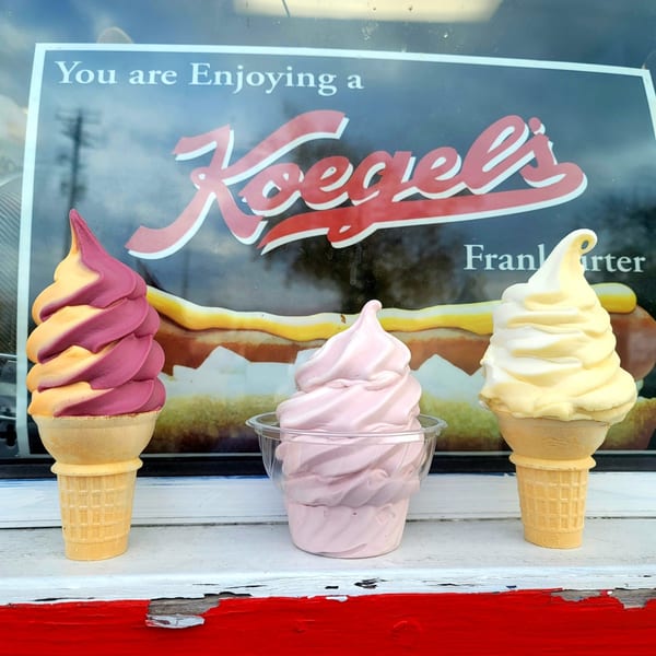 Three soft-serve ice creams, two in cones and one in a cup, in front of a Koegel's sign.